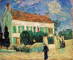 A white two-storey house at twilight, with two cypress trees on one end, and smaller green trees all around the house, with a yellow fence surrounding it. Two women are entering through the gate in the fence; a woman in black walks on by going towards the left. In the sky, there is a bright star with a large yellow halo around it